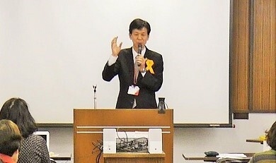 写真：胸にオレンジリボンを付けて挨拶をする区長（会議室内）