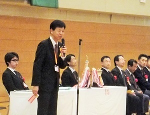 写真：胸にオレンジリボンを付けて挨拶する区長（講堂にて）