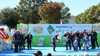 写真：舞台上に多くの男女が並んでいます。左側で何人かは歌を歌い、右側の8名程度の男女は紅白の幕でつつまれた木材を担いでいます。先頭には1人の男性が棒の先に白い紙をつけたものを振っています。3人の子供も一緒にステージに上がっています。