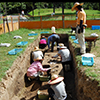 細長い調査区のなかで、参加者の民さんが遺構を掘り進めています。学芸員が全体の様子を見つめています。